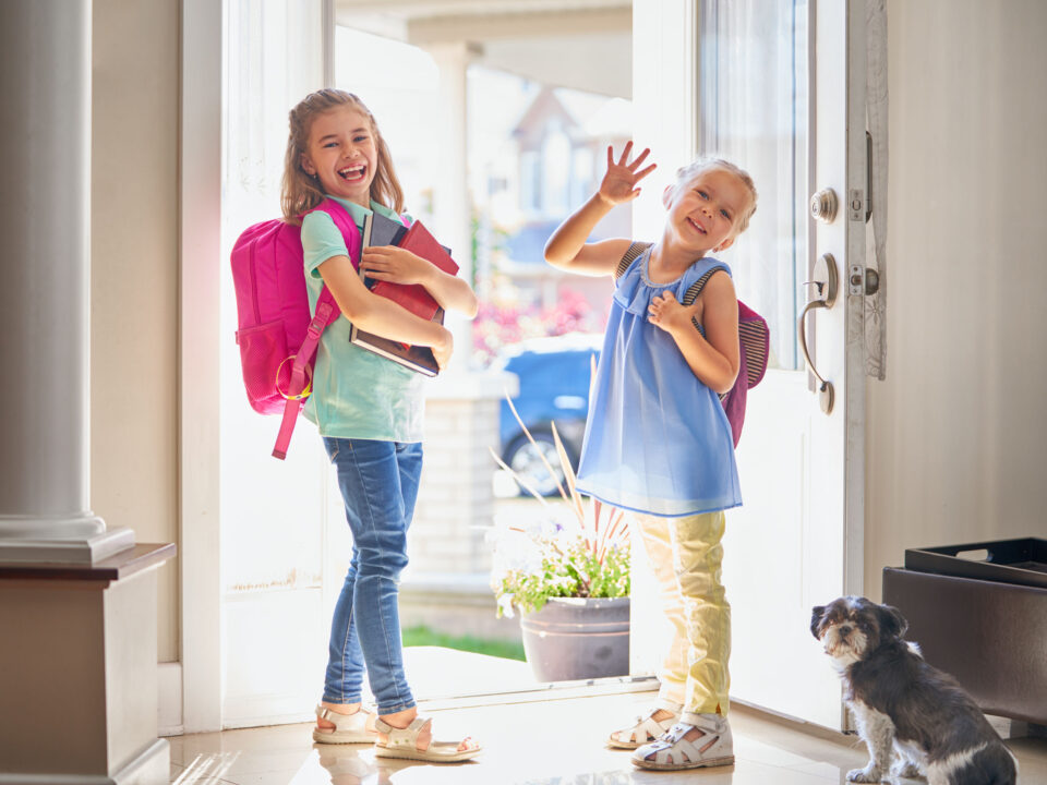 two girls with backpacks smiling and heading out the door to go to school