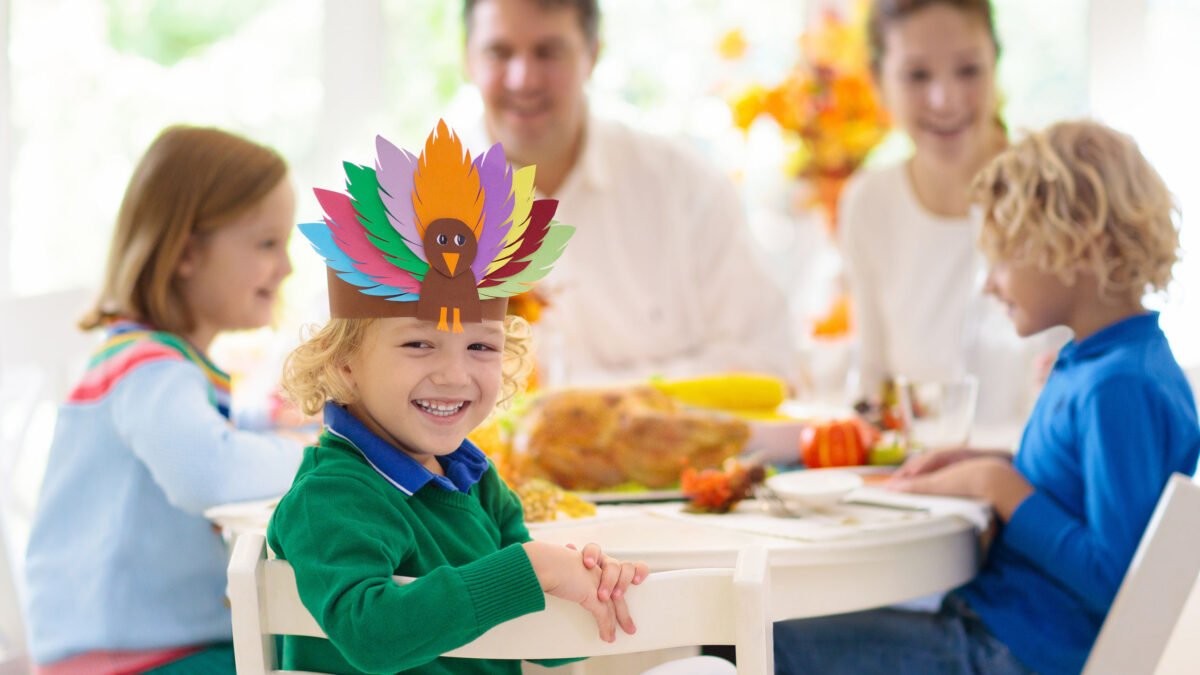 a little boy wearing a paper turkey headband and sitting around a table with his family on Thanksgiving