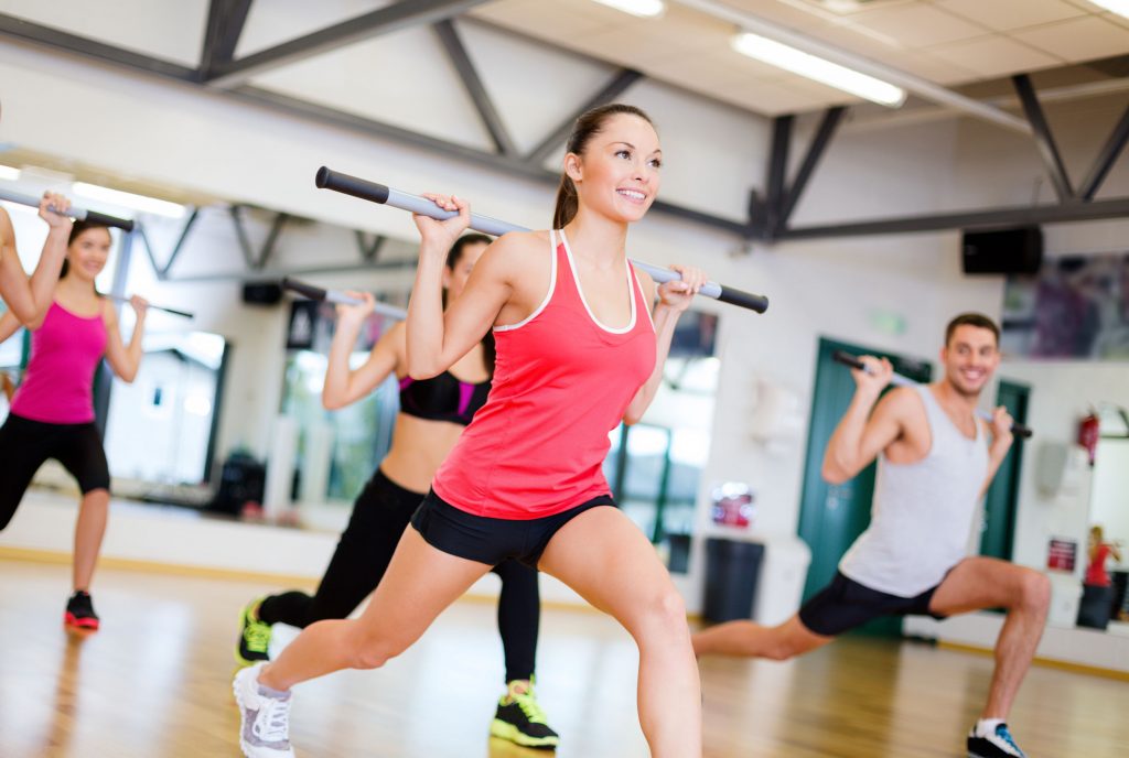 men and women smiling while lunging with a bar